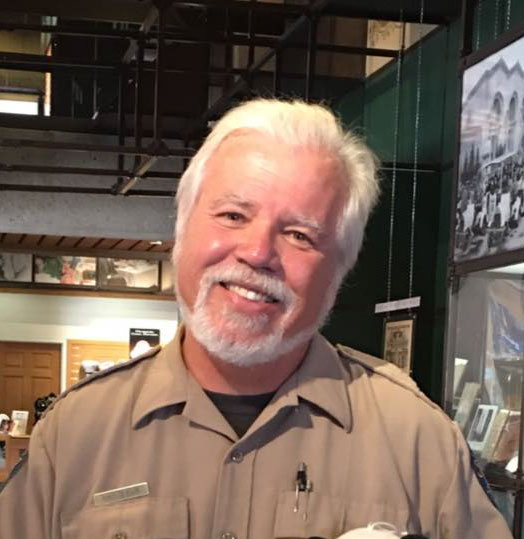 Photo of white man with white hair and beard wearing a brown shirt