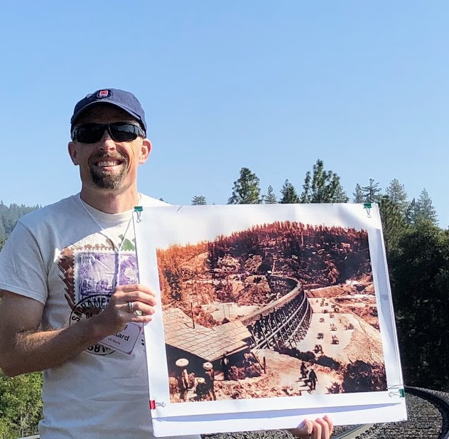 Photo of white man in the mountains holding an historical picture of a railroad trestle